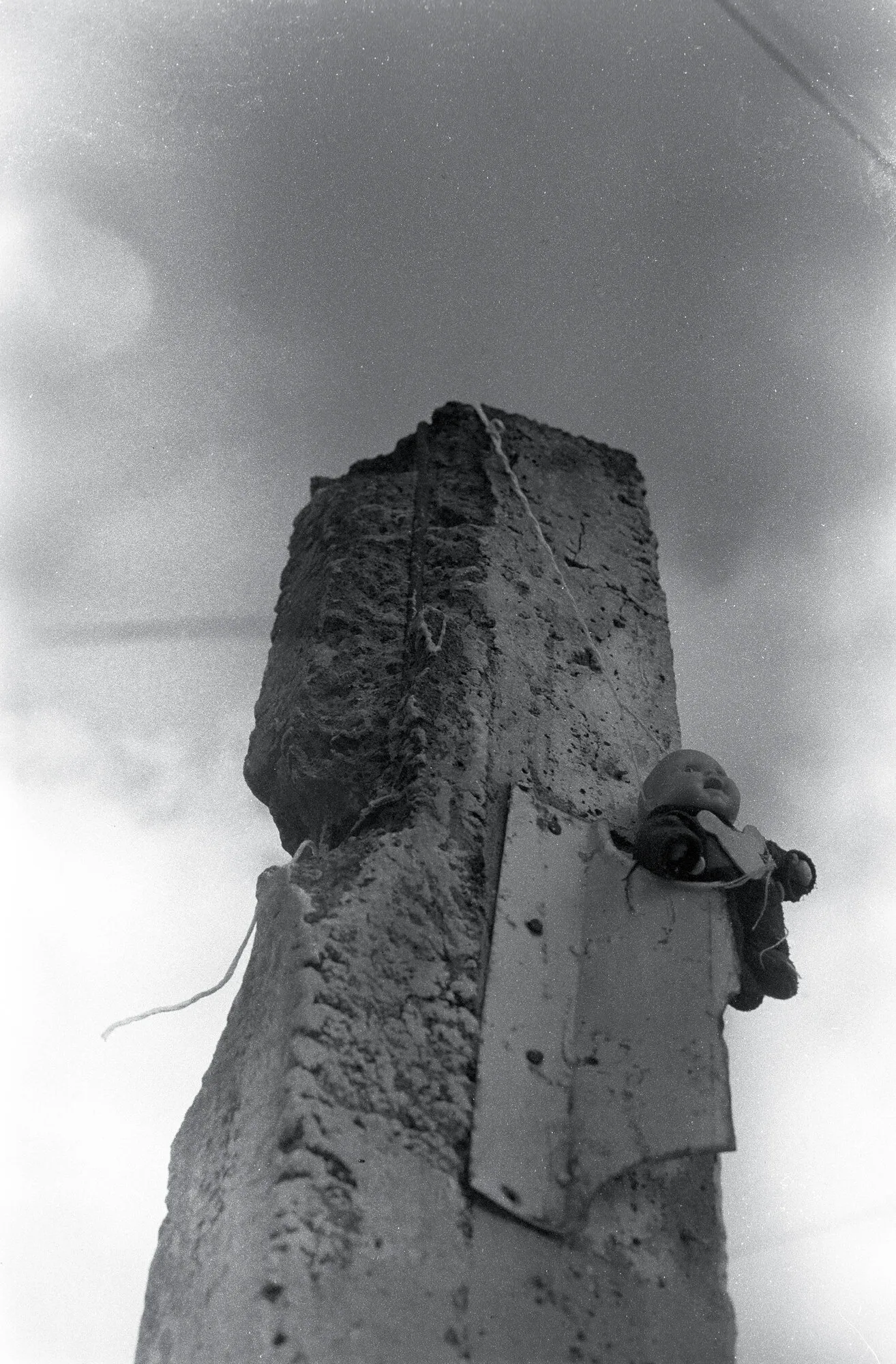 Un muñeco amarrado a lo alto de un poste de concreto roto, contra el cielo nublado, en blanco y negro.