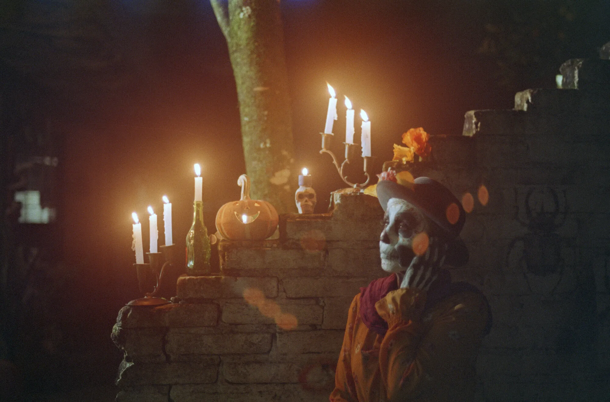 Una catrina con sombrero de flores mira hacia arriba junto a un muro con velas encendidas, una calabaza y calaveritas.