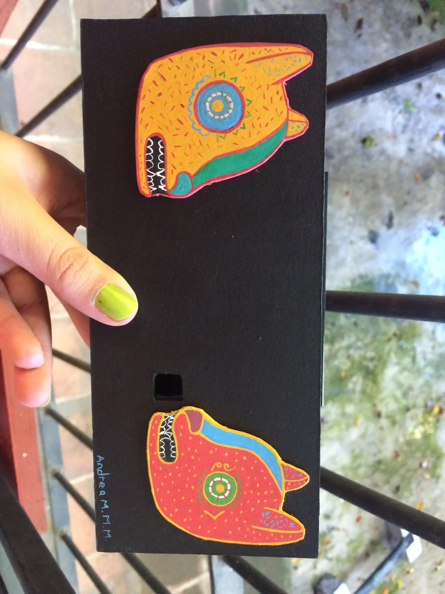 Cámara alargada de cartón negro con dos alebrijes pintados, sostenida en la mano
