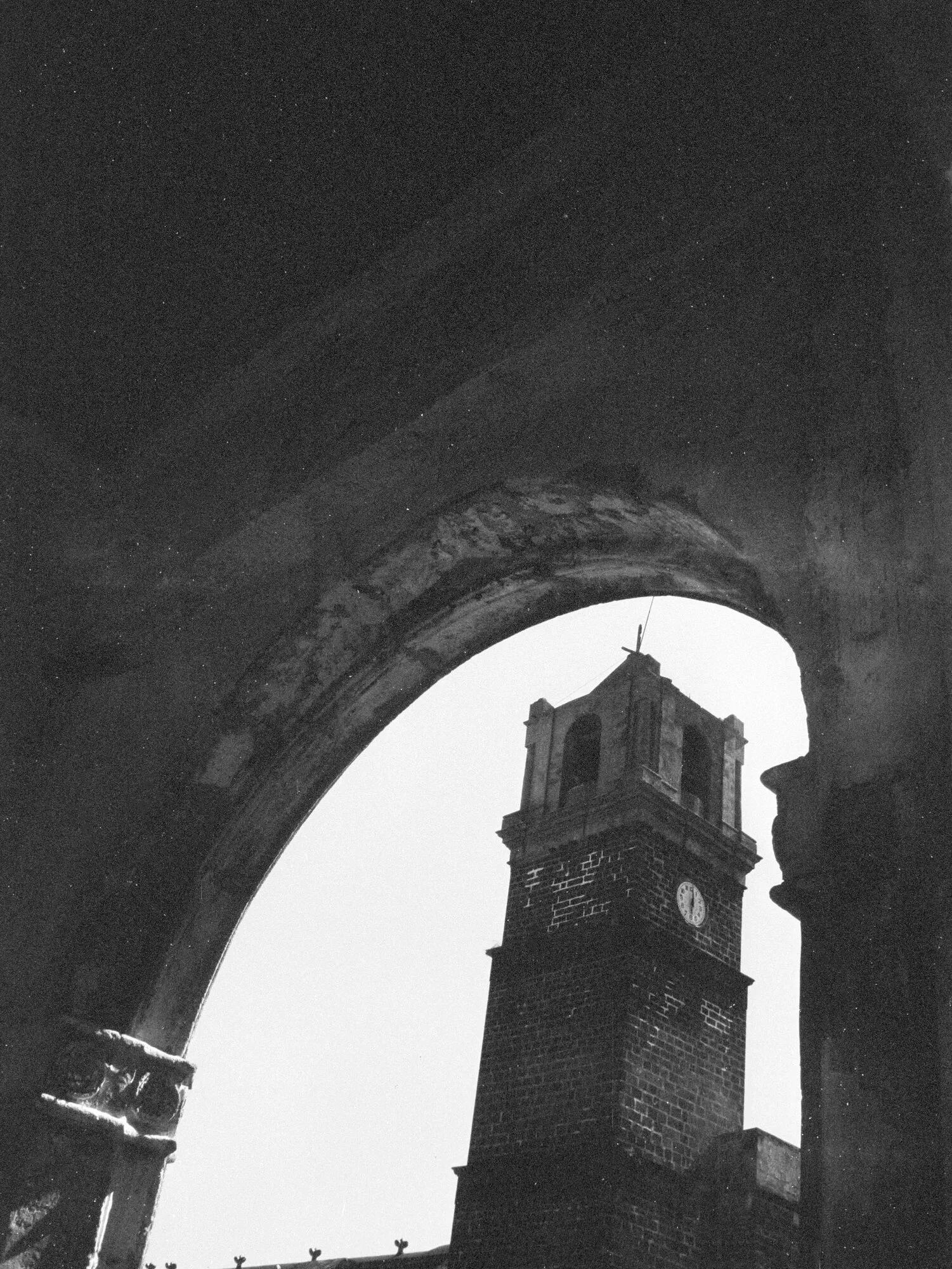 Torre de ladrillo con reloj vista a través de un arco de piedra, en blanco y negro.